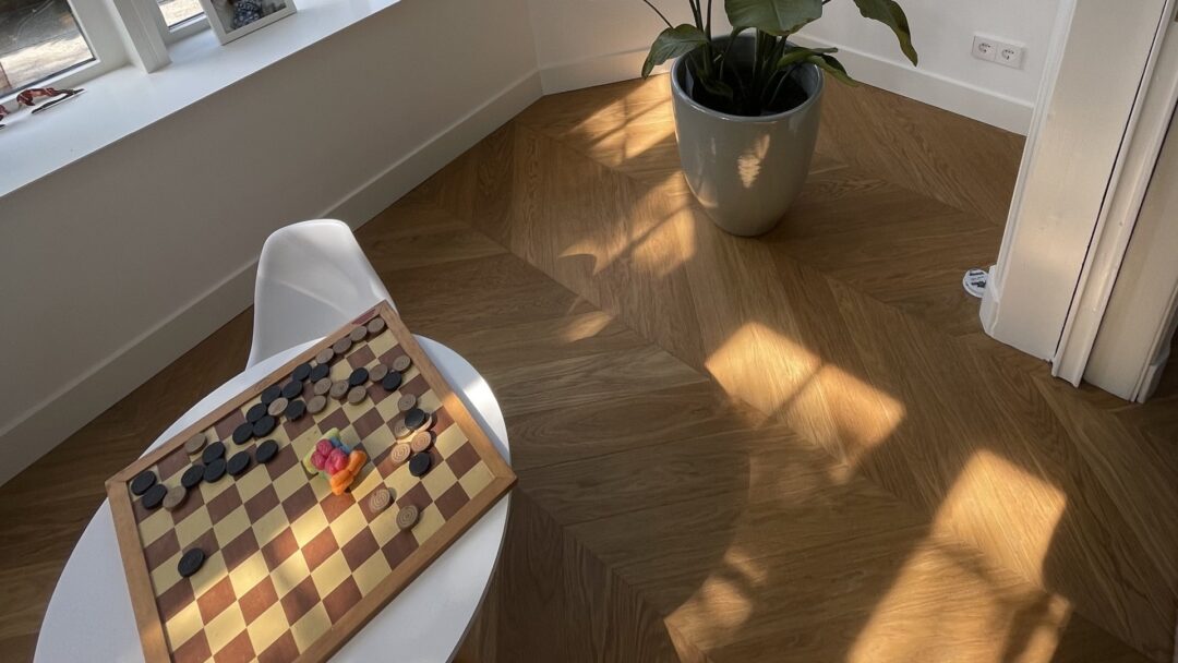 Hongaarse Punt Hoek van een interieur met een houten vloer in Hongaarse‑punt patroon gelegd door Themen Floors, een plant in een grote pot en een rond wit tafeltje en stoel met een dambord en speelstukken.
